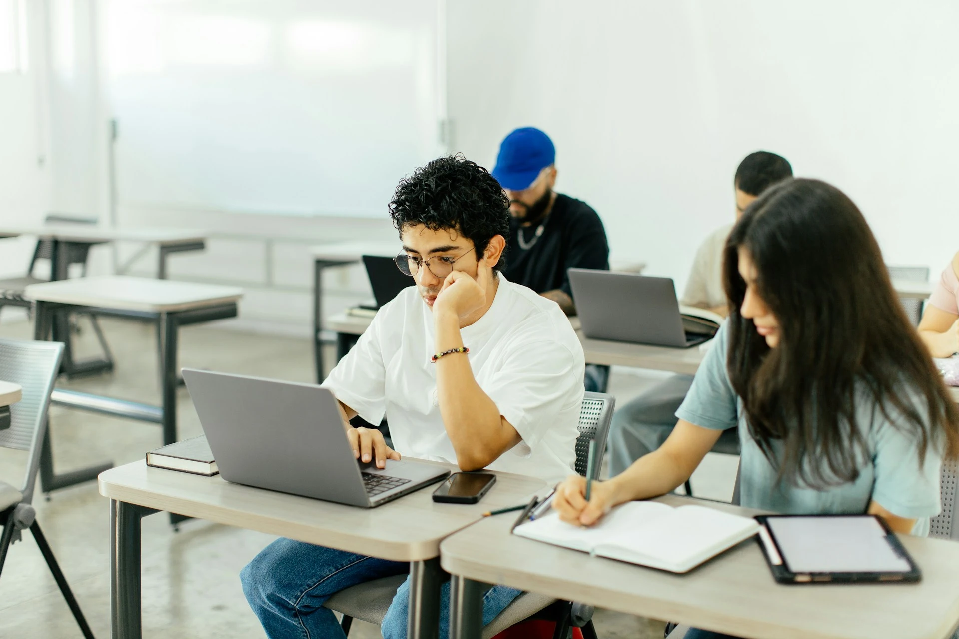 Students sitting at desks working on laptops and taking notes in a bright classroom.